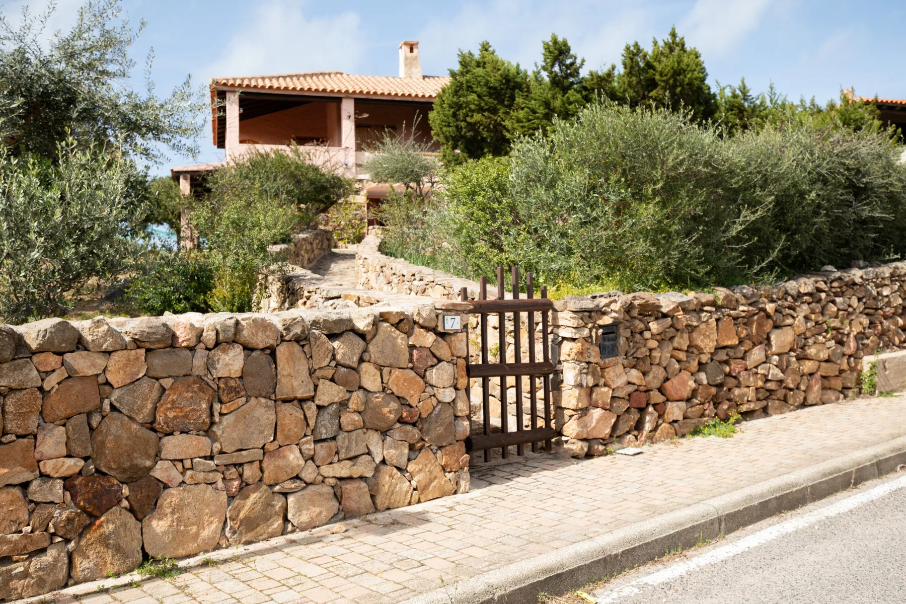 A stone wall and wooden gate line a paved walkway in front of a house.