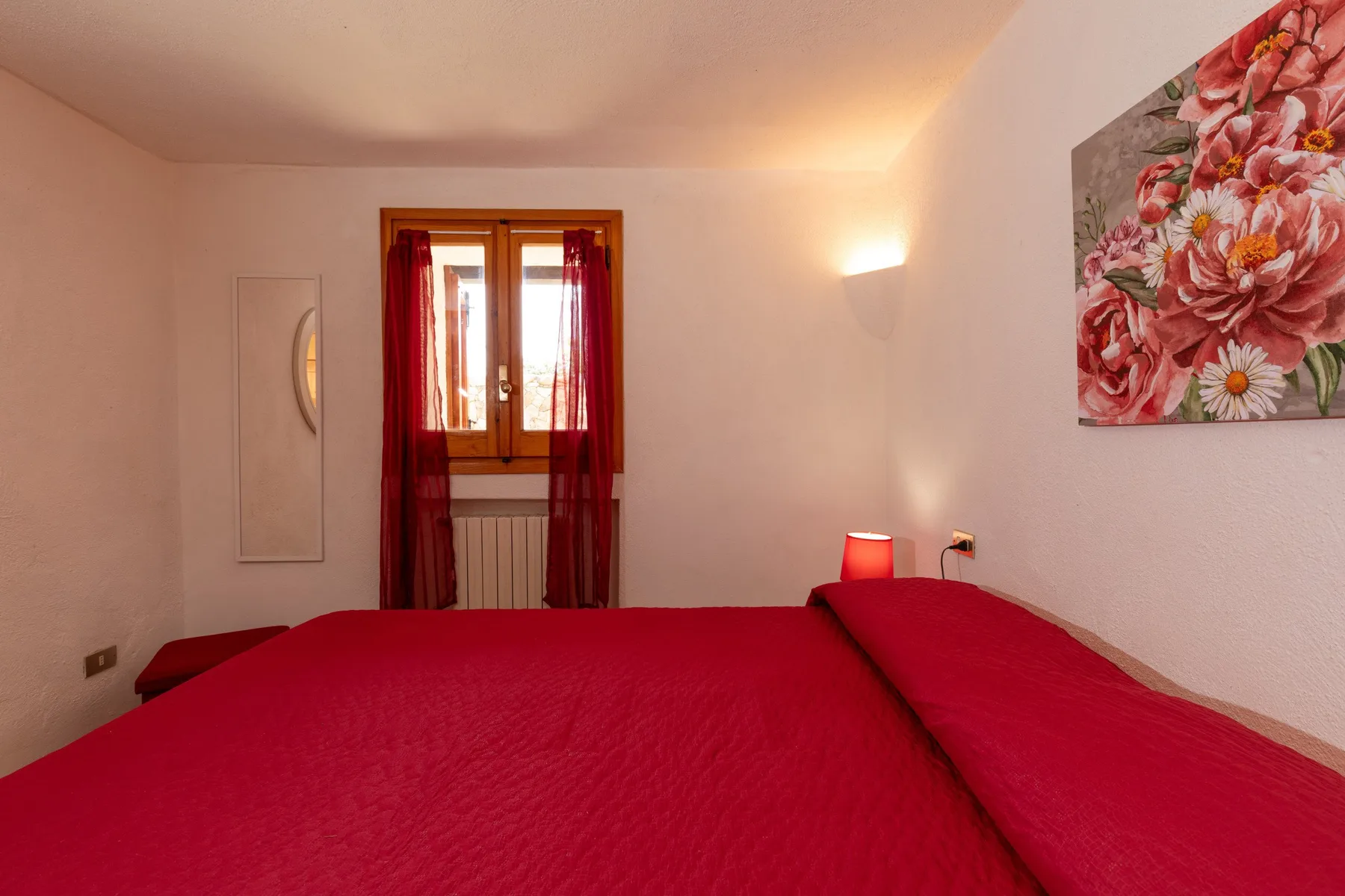 Un dormitorio con ropa de cama roja, una ventana y un cuadro floral.