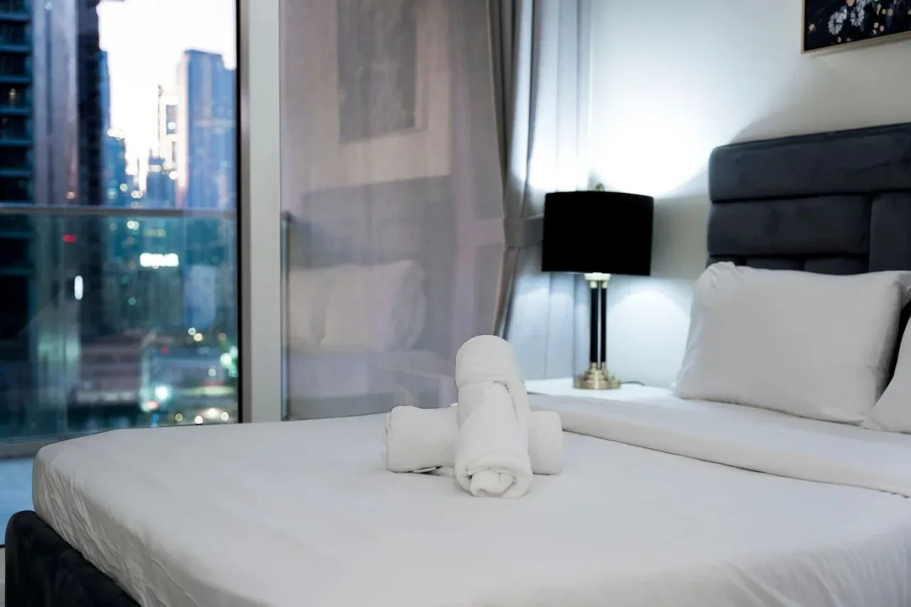 Dormitorio con una cama bien arreglada y una ventana con vistas a edificios cercanos.