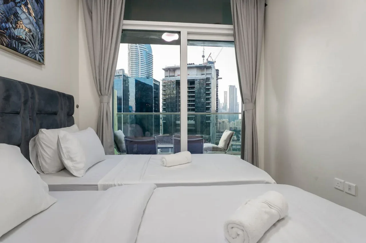 Un dormitorio con camas blancas y una ventana grande con vista a edificios cercanos.