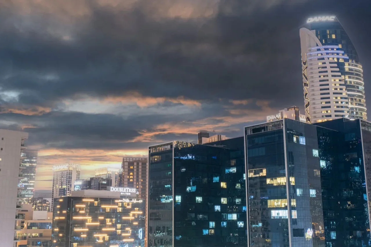 Los edificios de la ciudad brillan bajo un cielo nublado al anochecer.