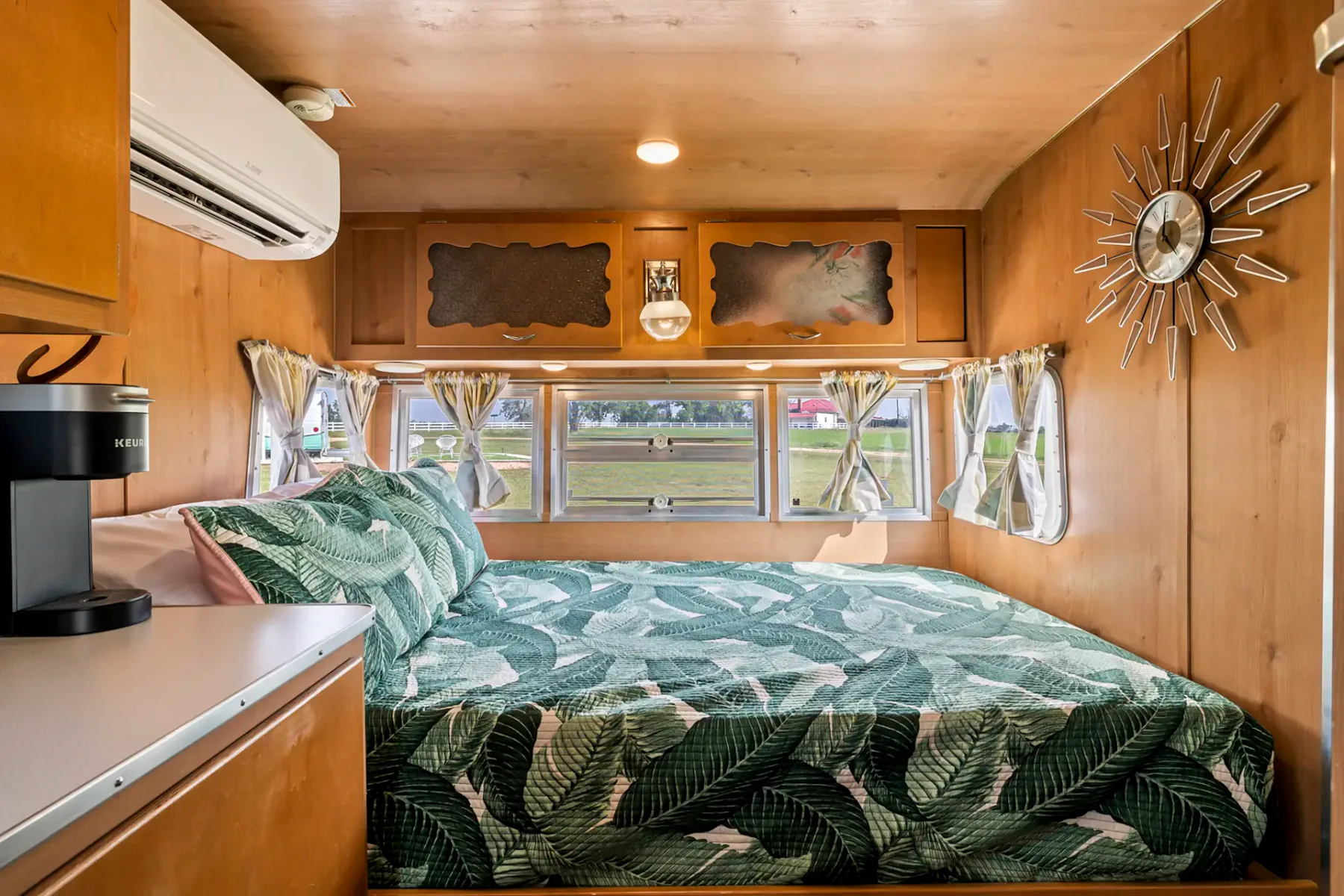 Acogedor dormitorio de caravana con una colcha estampada con hojas tropicales, paredes de madera, ventanas con cortinas florales y un reloj antiguo en la pared.