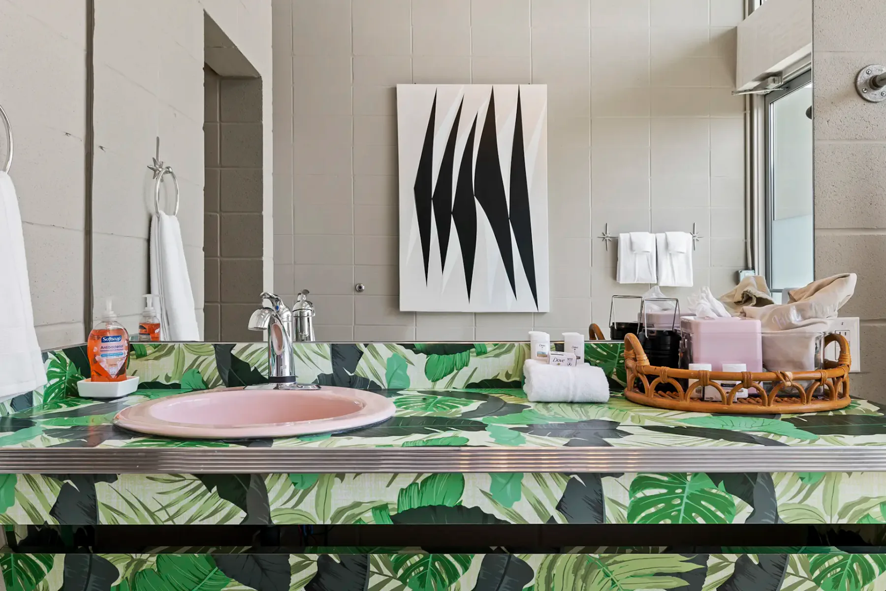 Baño moderno con grandes ventanas, mueble de lavabo verde estampado, espejo, albornoces colgados y una cesta tejida sobre el suelo de hormigón.