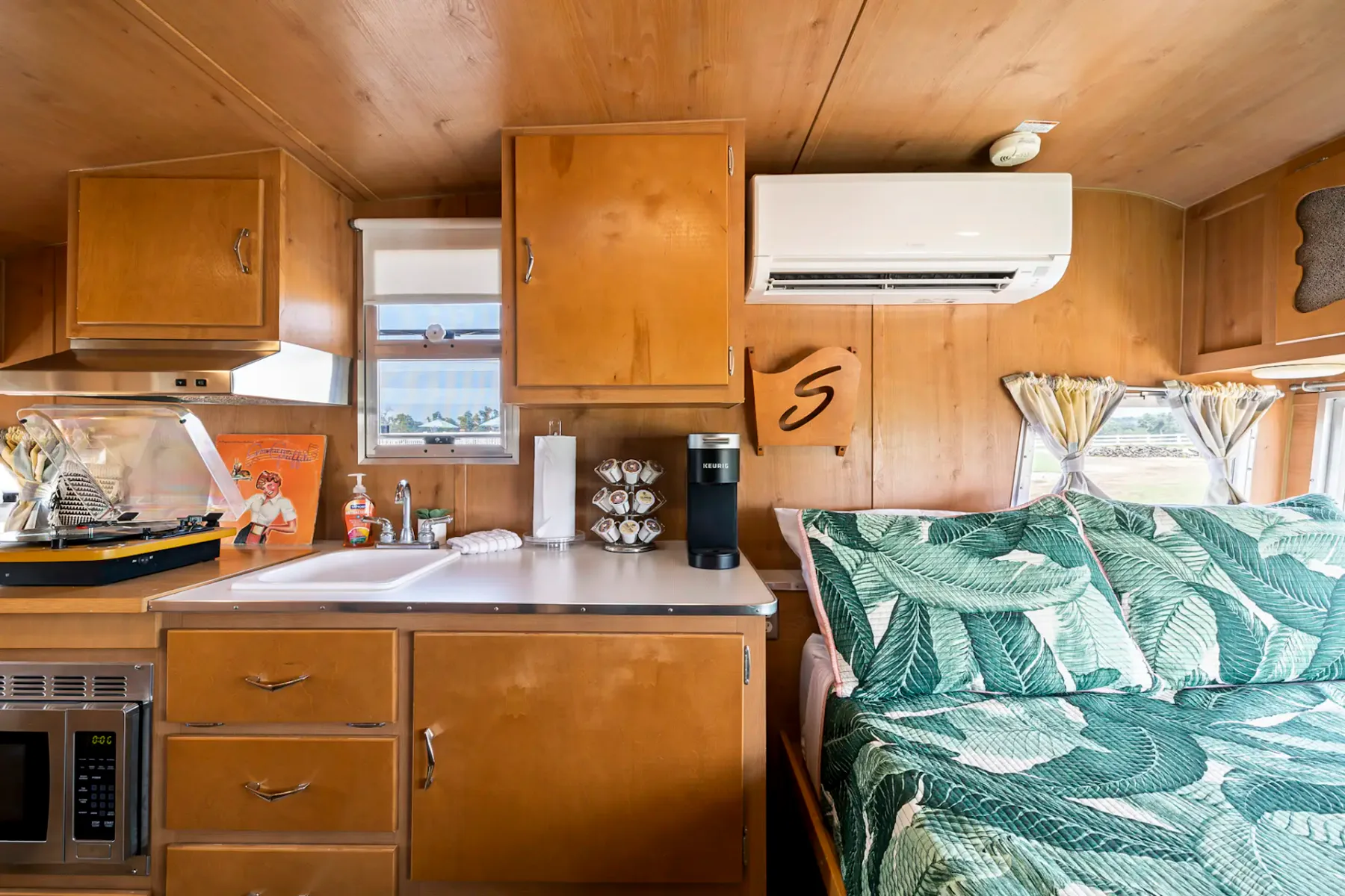 Acogedor dormitorio de caravana con una colcha estampada con hojas tropicales, paredes de madera, ventanas con cortinas florales y un reloj antiguo en la pared.