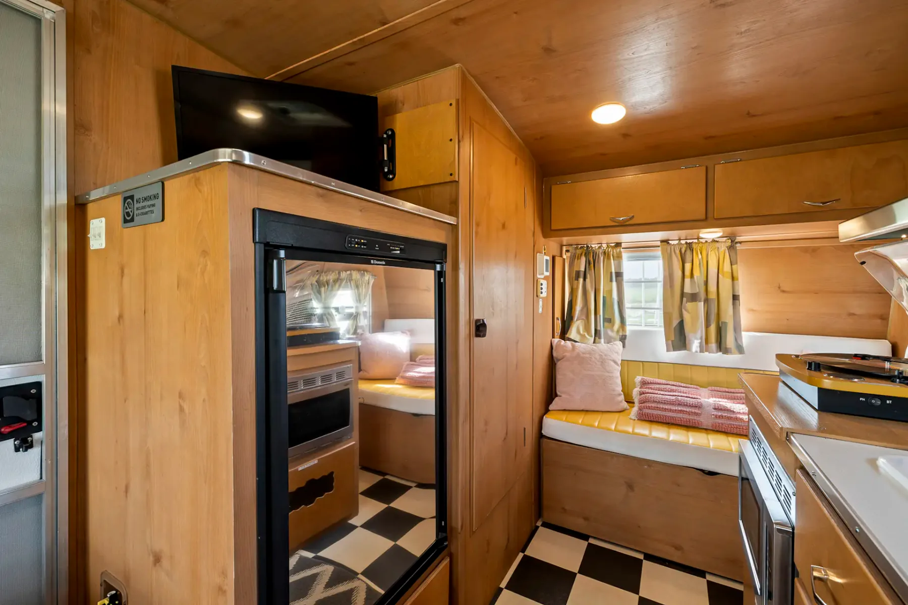 Acogedor interior de caravana de madera con cocina pequeña, nevera pequeña y zona de estar. Suelo ajedrezado en blanco y negro, televisor y cojines de colores.
