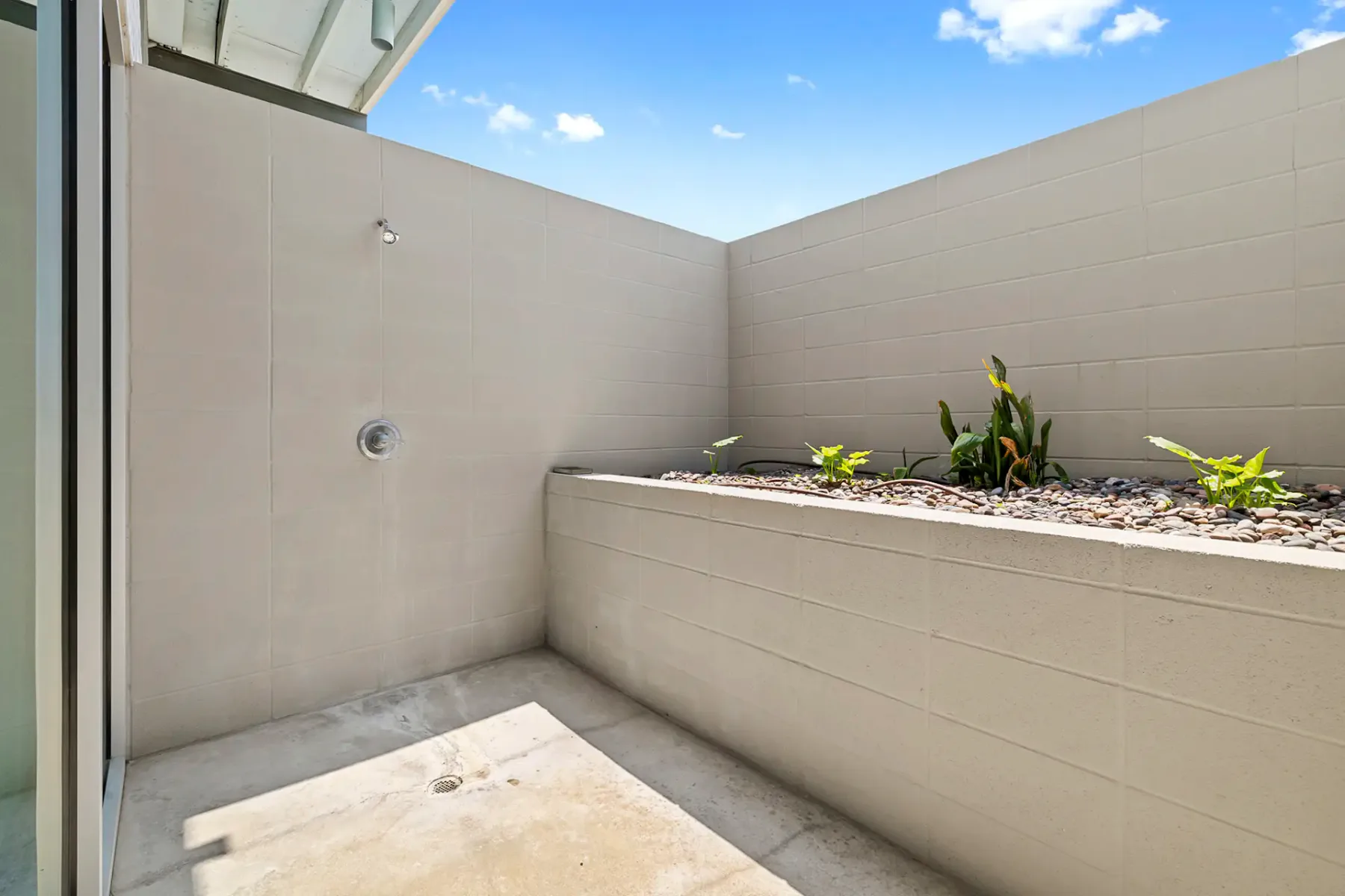 Zona de ducha al aire libre con paredes revestidas de azulejos beige, desagüe en el suelo, cabezal de ducha montado en la pared y una jardinera elevada con plantas pequeñas. Arriba se ve el cielo azul.