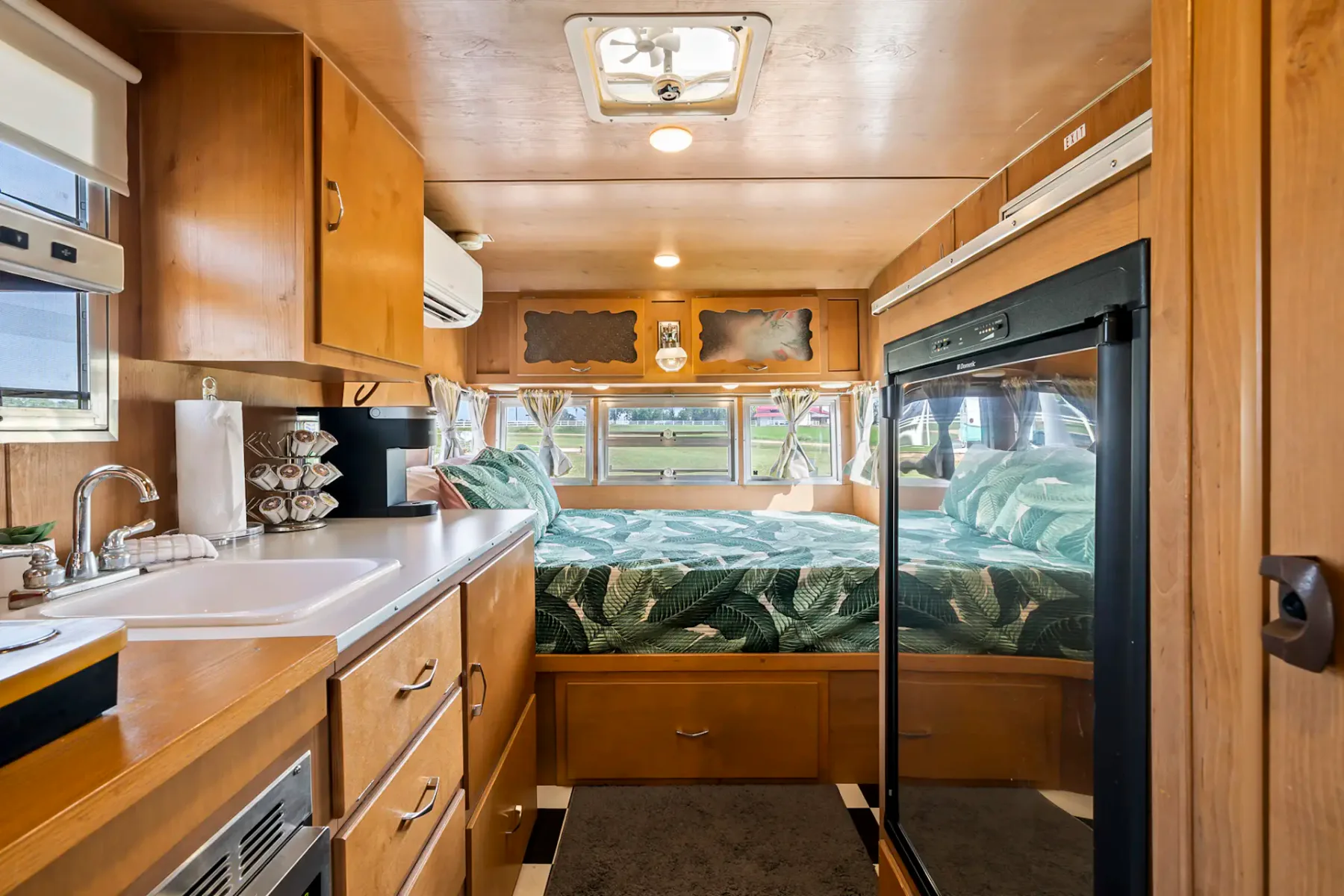 Interior de una acogedora autocaravana con cocina pequeña, fregadero y cama. Los armarios de madera, las grandes ventanas y los electrodomésticos compactos crean un espacio acogedor.