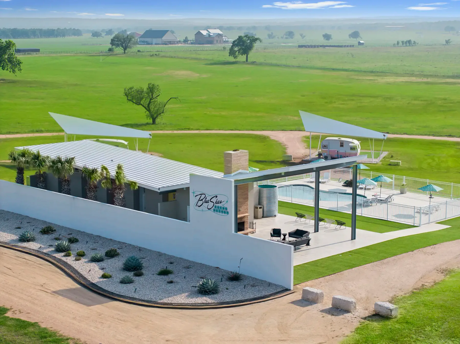 Casa moderna de una planta con patio, piscina y caravana vintage en un extenso campo verde. Al fondo se ven edificios agrícolas lejanos y algunos árboles.