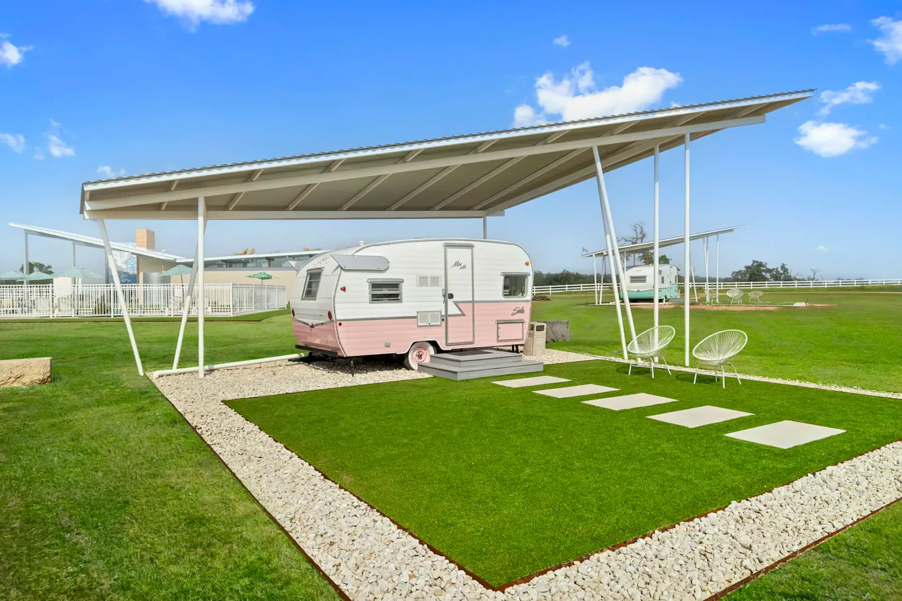 Una caravana vintage rosa y blanca está estacionada sobre el césped bajo una gran cubierta metálica angular, con losas de piedra y bordes de guijarros blancos.