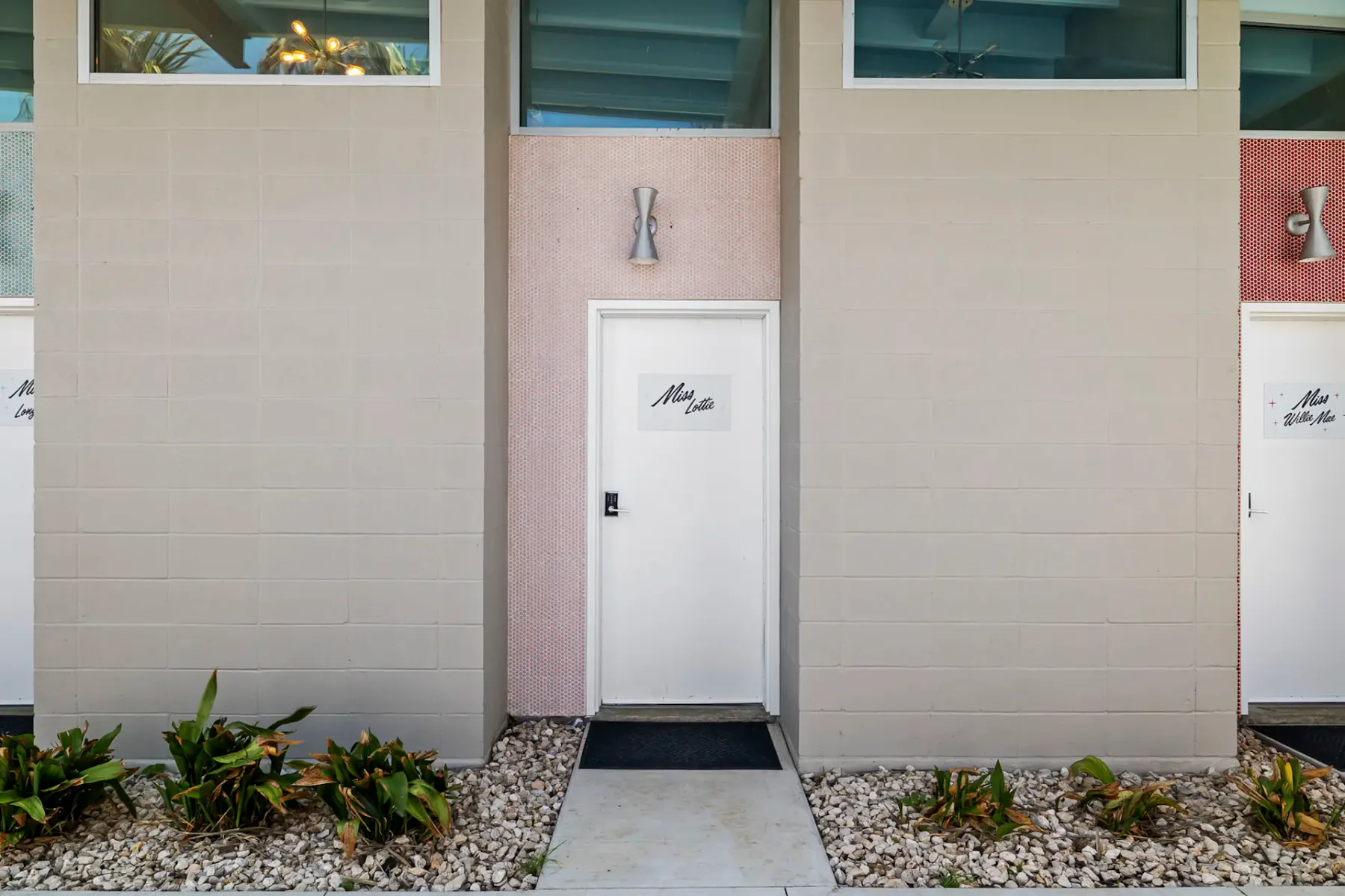 Fachada de edificio minimalista con puerta blanca, plantas pequeñas, suelo de guijarros y paredes rosas y beige, con apliques de pared modernos.
