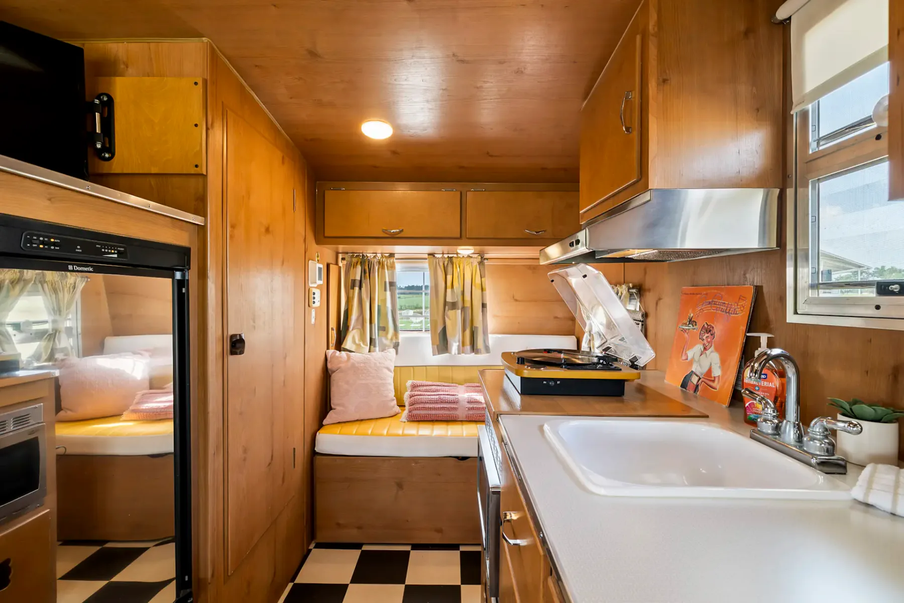 Cocina retro con armarios de madera, un fregadero pequeño, cafetera, microondas, tocadiscos y un portarrollos de papel de cocina cerca de una ventana pequeña.