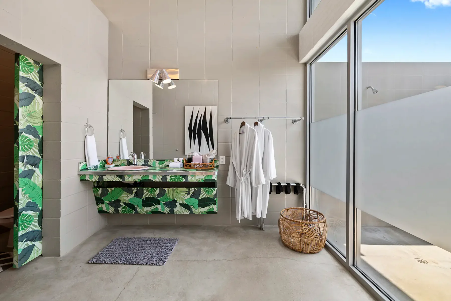 Baño moderno con grandes ventanas, mueble de lavabo verde estampado, espejo, albornoces colgados y una cesta tejida sobre el suelo de hormigón.