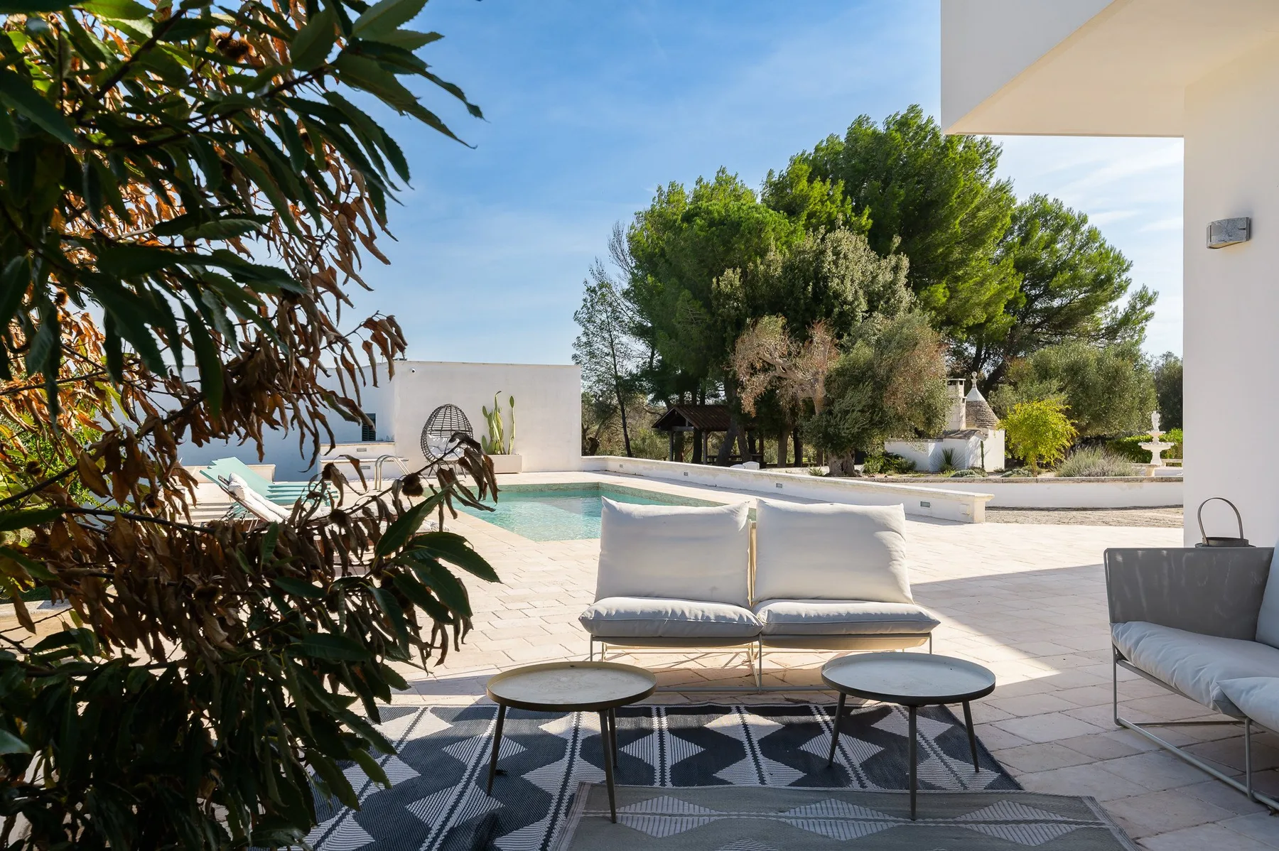 A sunlit poolside patio with outdoor seating and leafy trees.