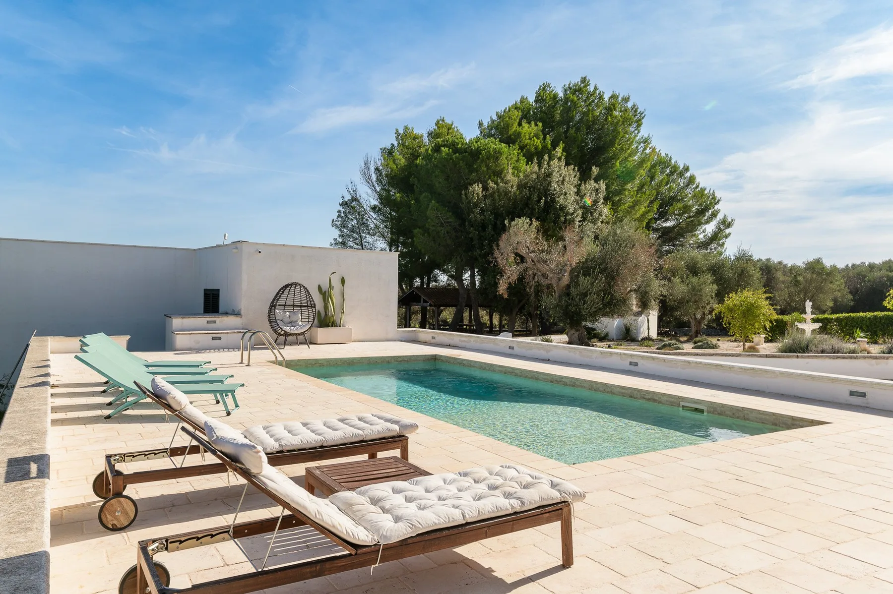 An outdoor pool beside a paved patio with lounge seating and trees.