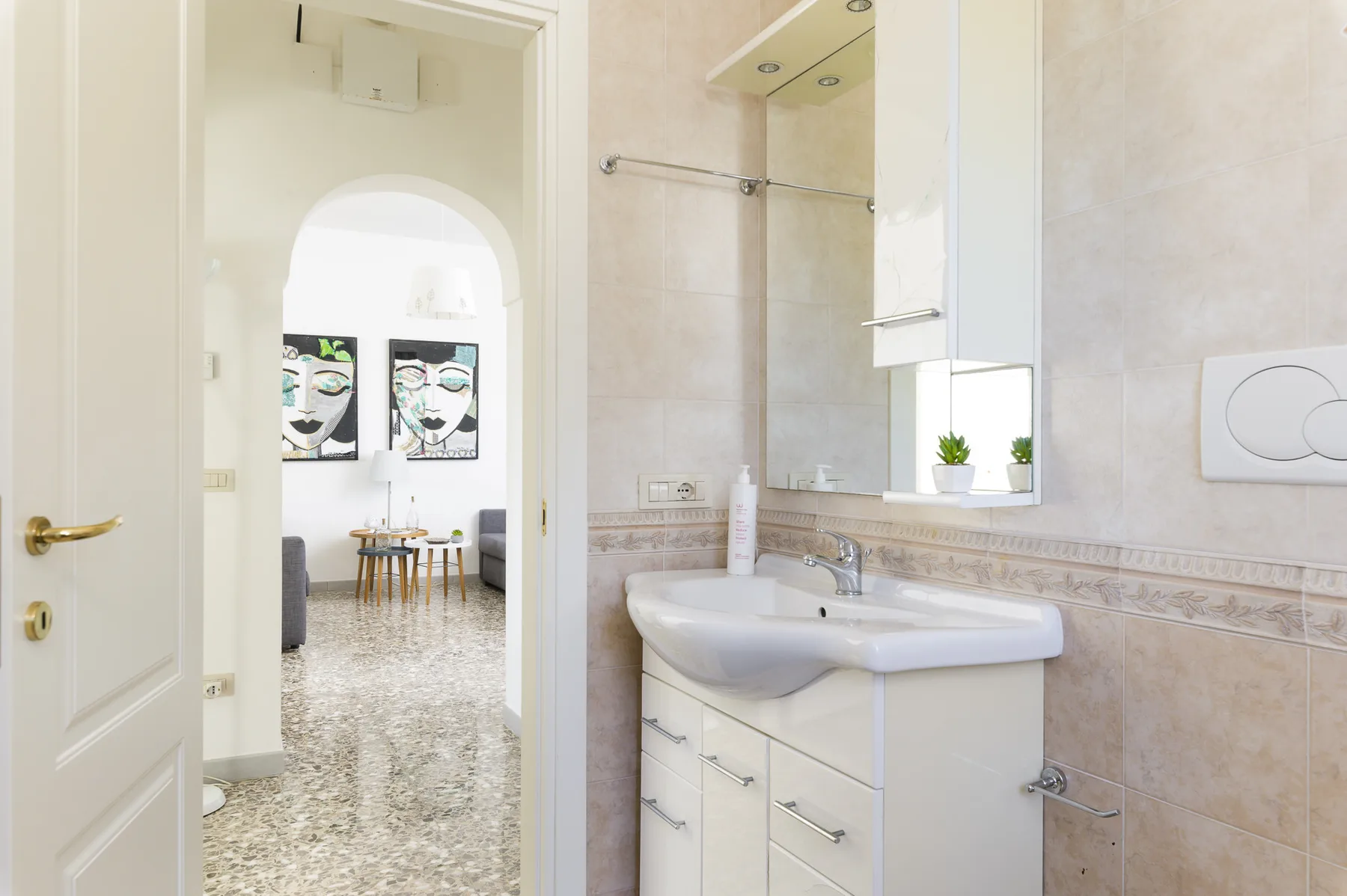 A tiled bathroom with a sink vanity and a doorway opening onto a bright adjoining room.
