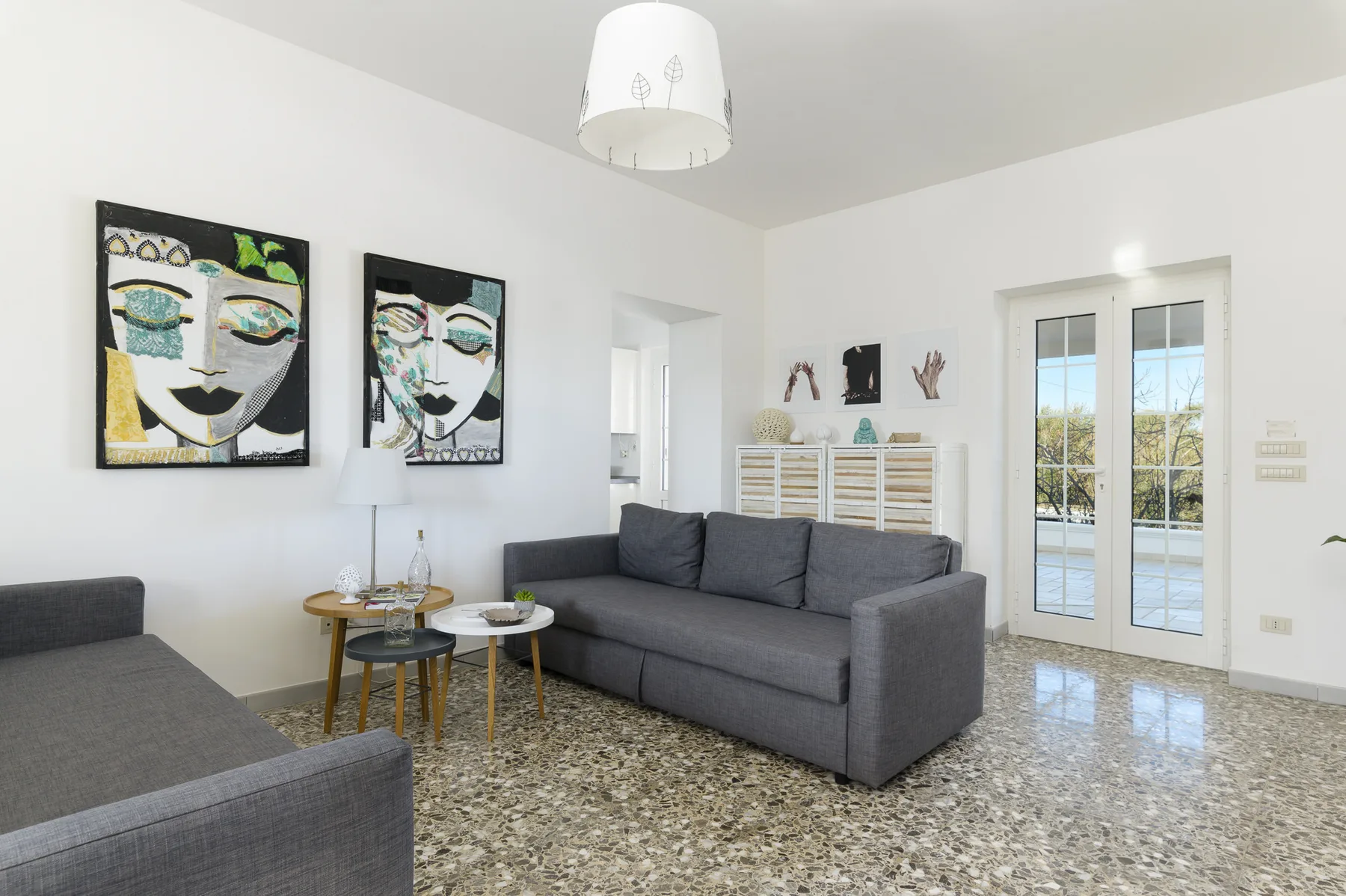 A bright living room with gray seating, colorful wall art, and glass doors.