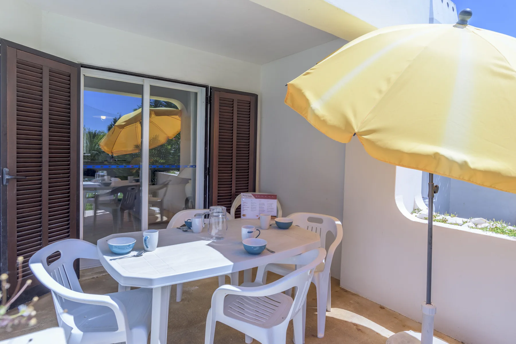 A covered patio dining area with a white table, plastic seating, and a yellow umbrella.