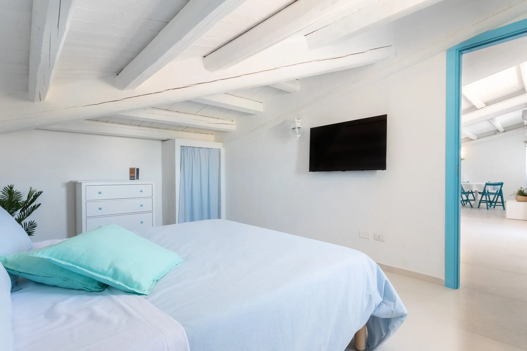 A bright bedroom with white bedding, a turquoise pillow, and a wall-mounted television.