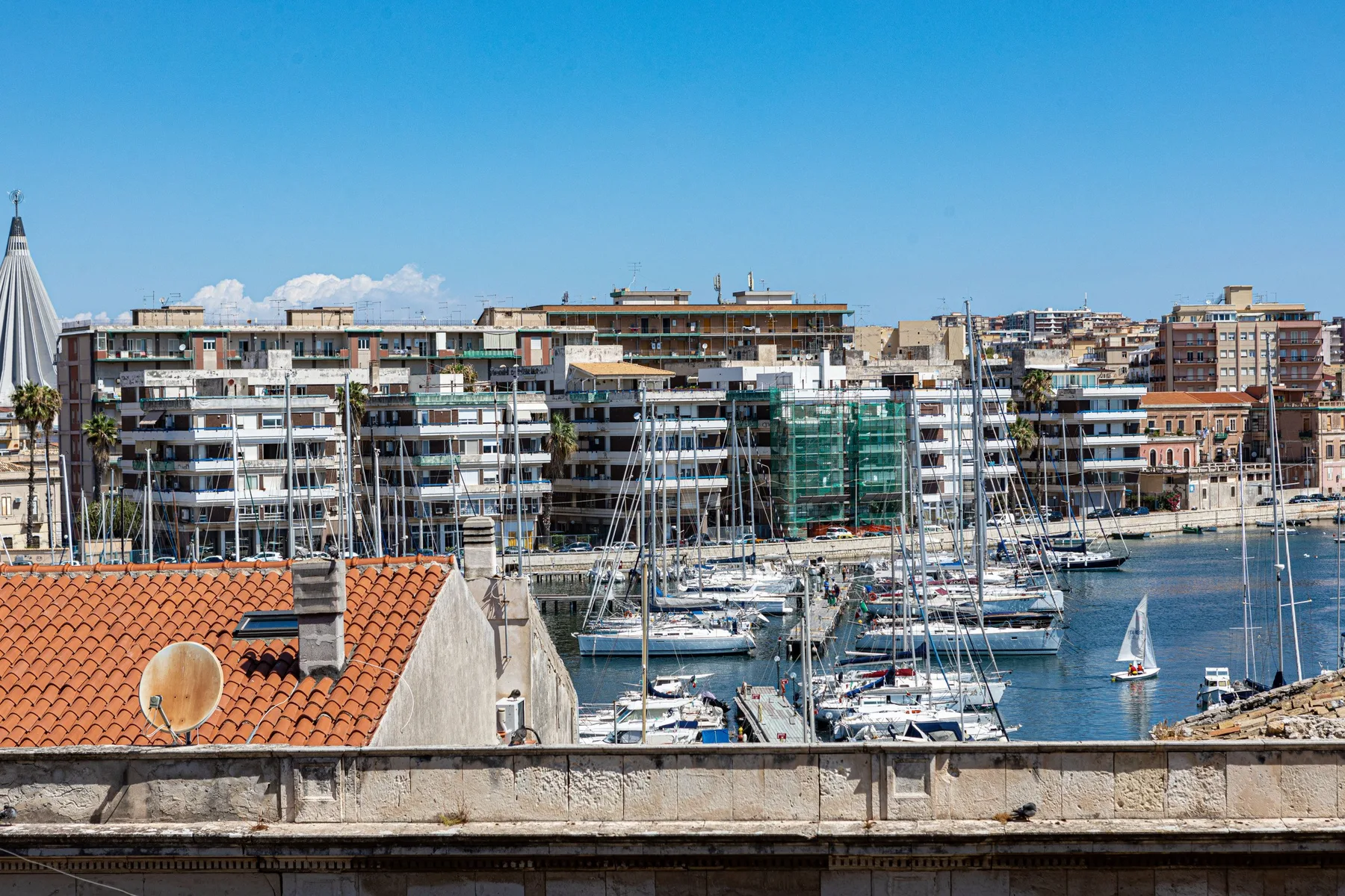 A marina filled with sailboats sits beside a waterfront lined with apartment buildings.