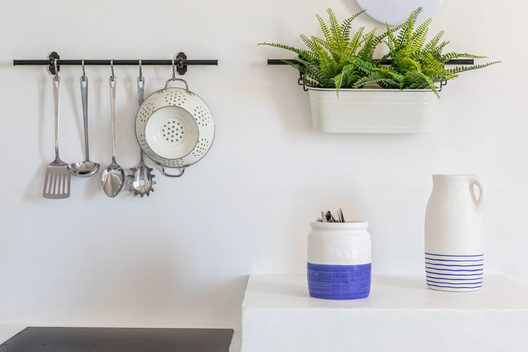 A bright kitchen wall has hanging utensils, a potted fern, and ceramic containers.