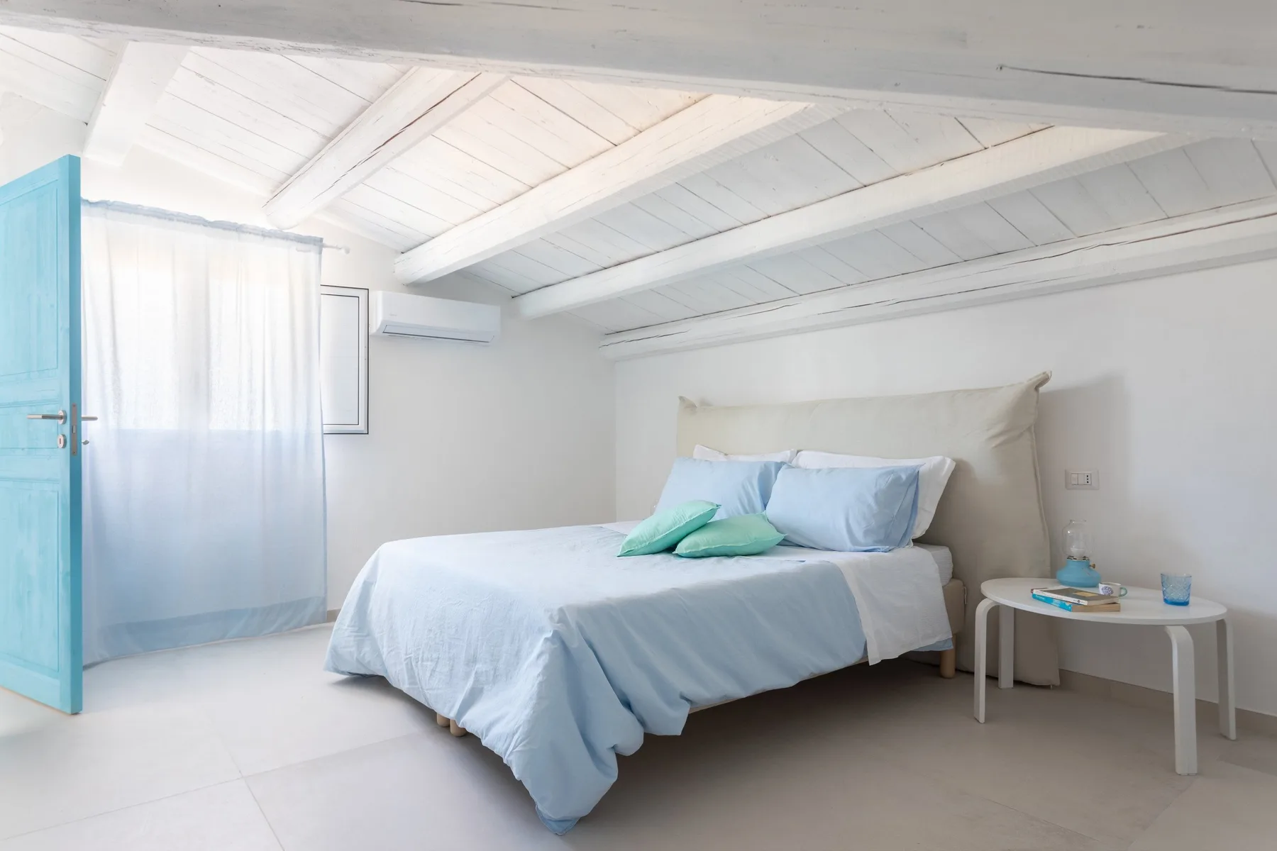 A bright bedroom with a bed dressed in pale blue linens and a small table.