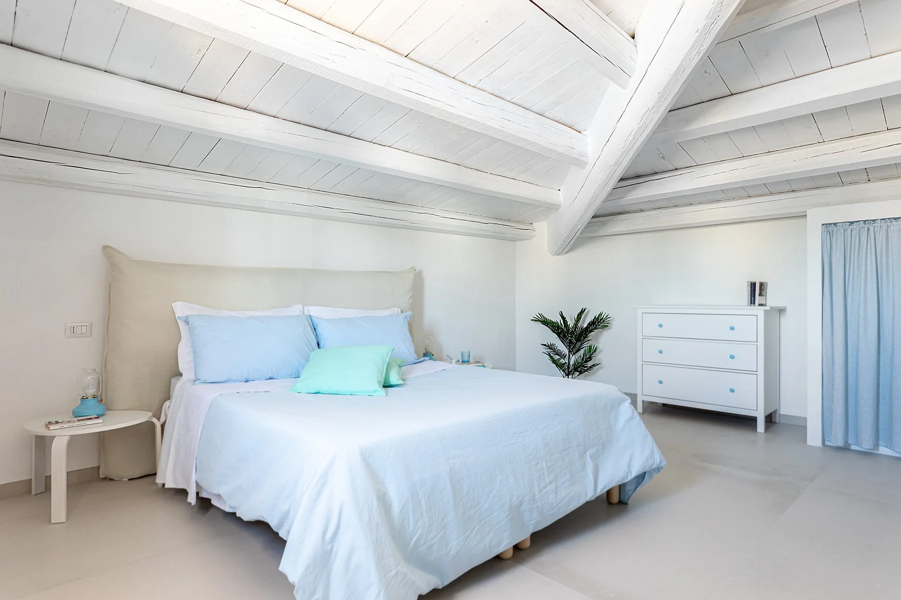 A bright bedroom with a neatly made bed beneath white exposed ceiling beams.