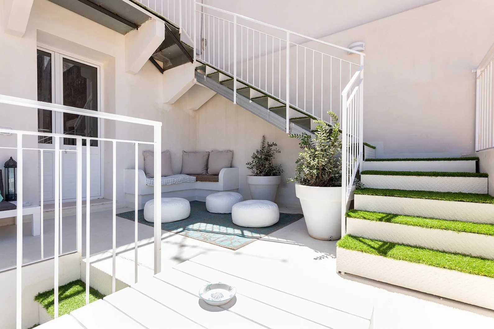 A bright outdoor patio has cushioned seating, potted plants, and steps edged with green turf.
