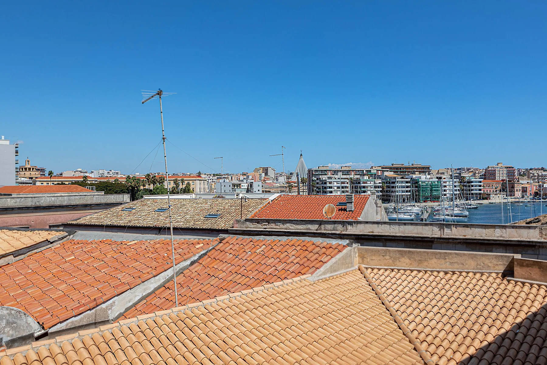 A city skyline rises beyond rows of terracotta rooftops under a clear blue sky.