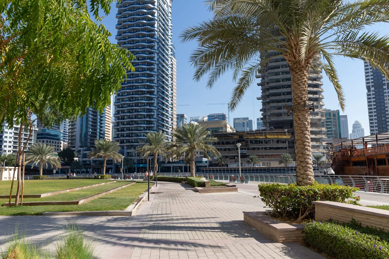 Un sendero pavimentado atraviesa una plaza urbana ajardinada con palmeras y edificios altos.