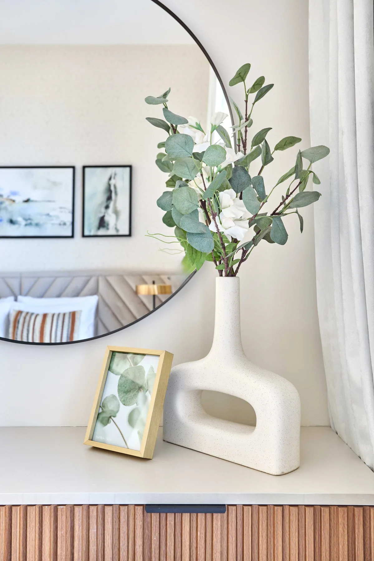 Un jarrón decorativo con ramas frondosas está sobre un mueble junto a un espejo redondo.