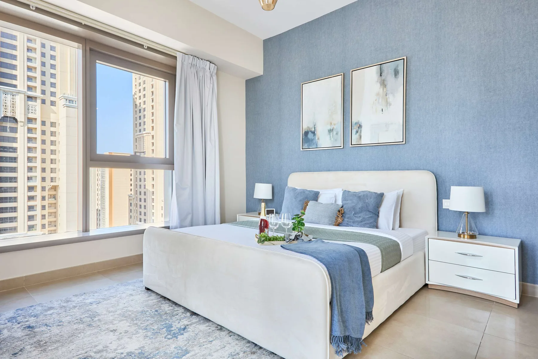Un dormitorio con una cama bien arreglada junto a grandes ventanas con vistas a edificios cercanos.