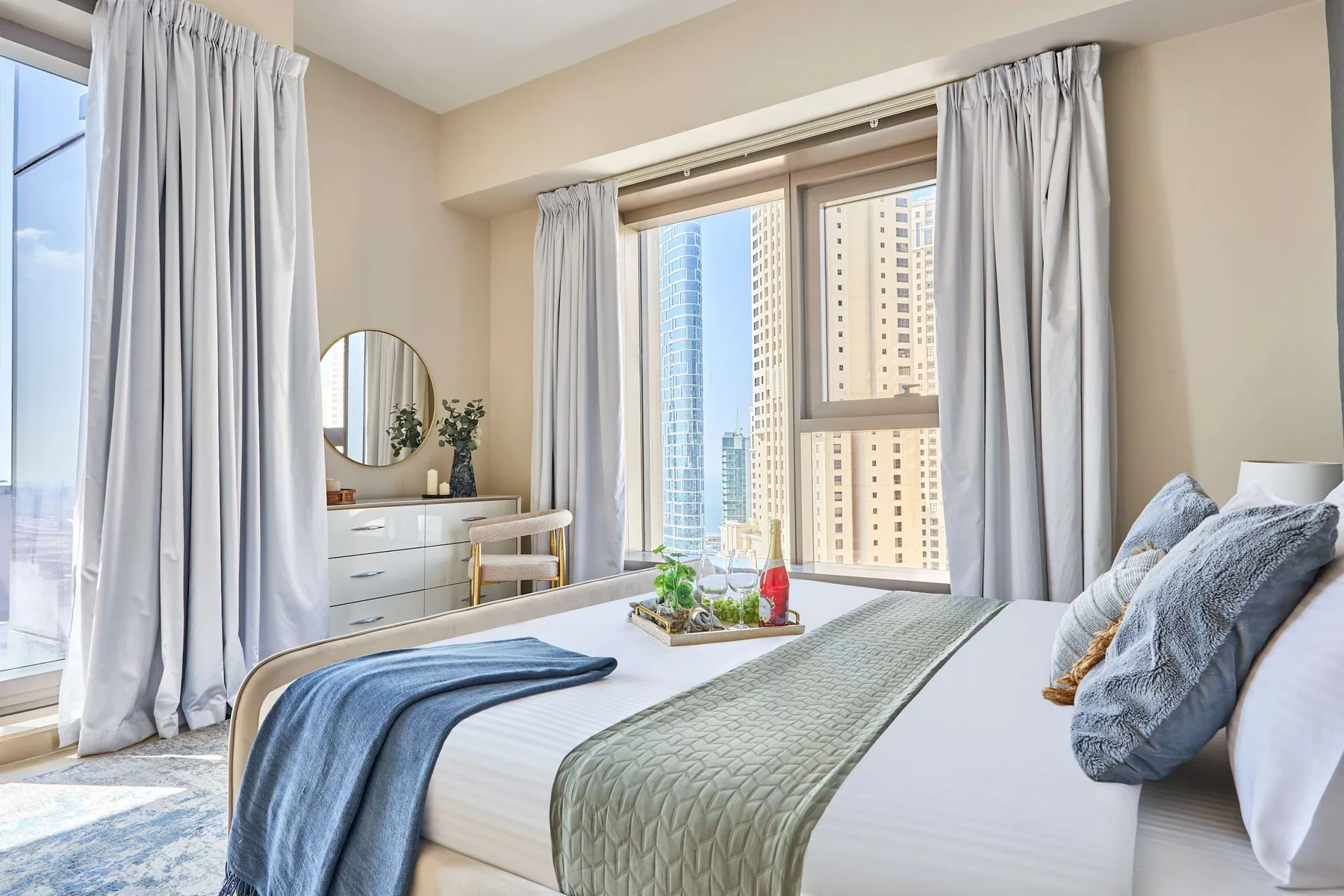 Un dormitorio con una cama bien tendida junto a grandes ventanas y cortinas claras.