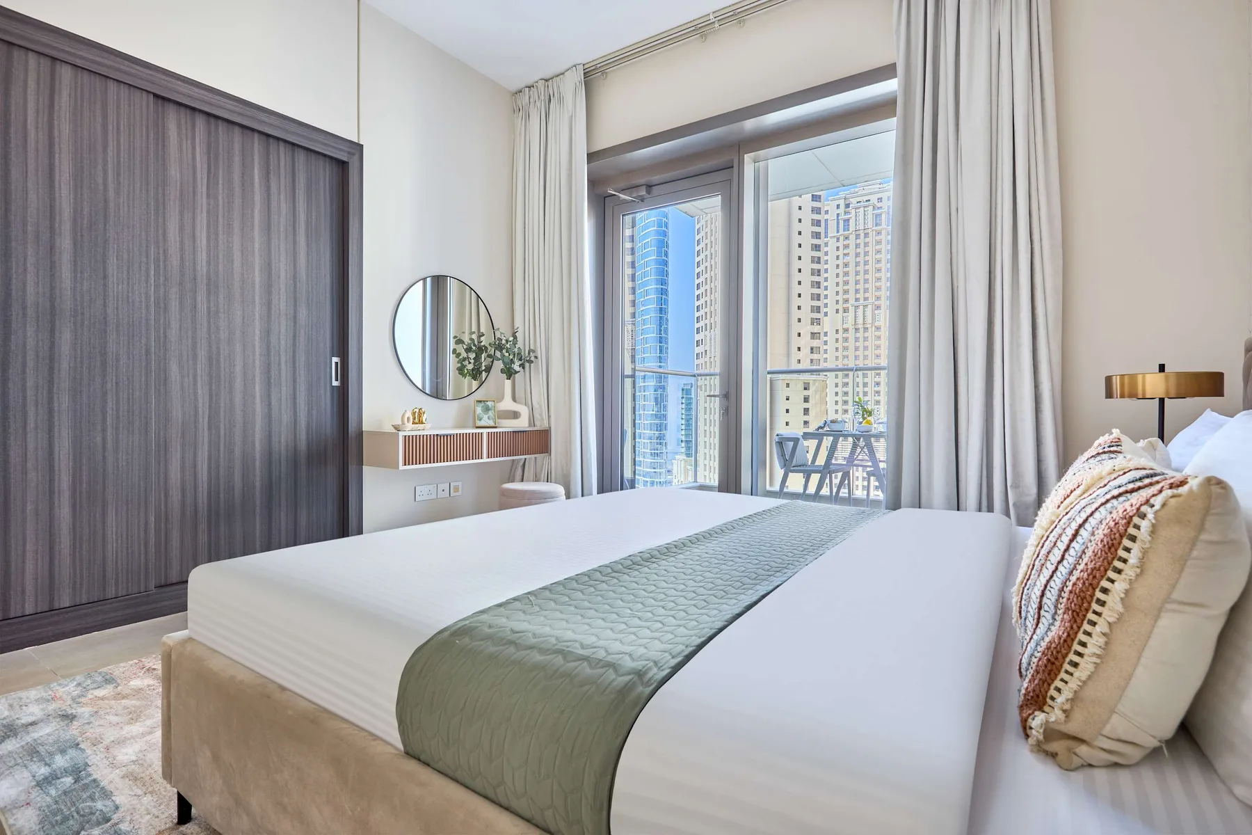 Un dormitorio luminoso con una cama, cortinas y grandes ventanas frente a edificios cercanos.