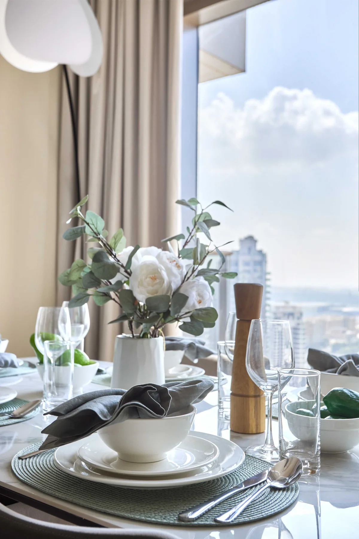 Una mesa de comedor está puesta con platos, copas y flores junto a una ventana grande.