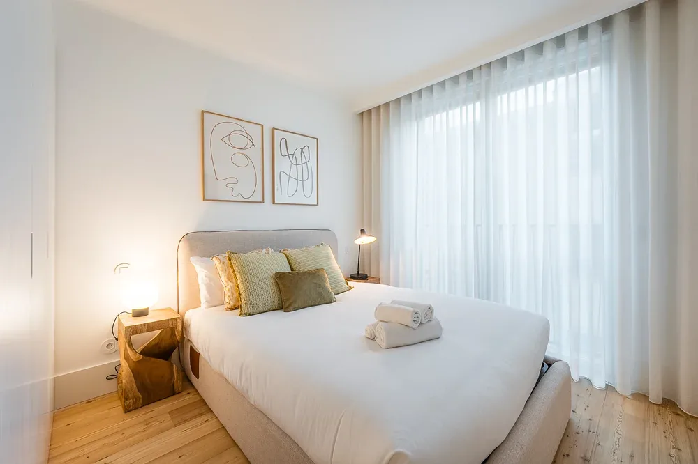 A bright bedroom with a neatly made bed beside a large curtained window.