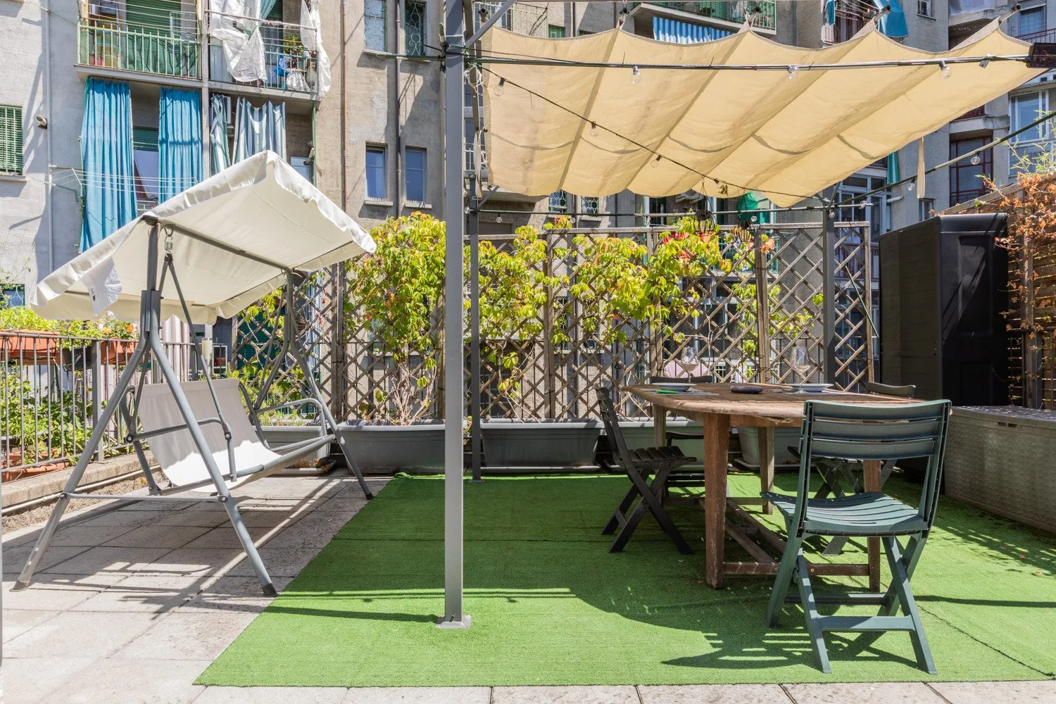 An outdoor terrace has green flooring, shaded seating, and a table.
