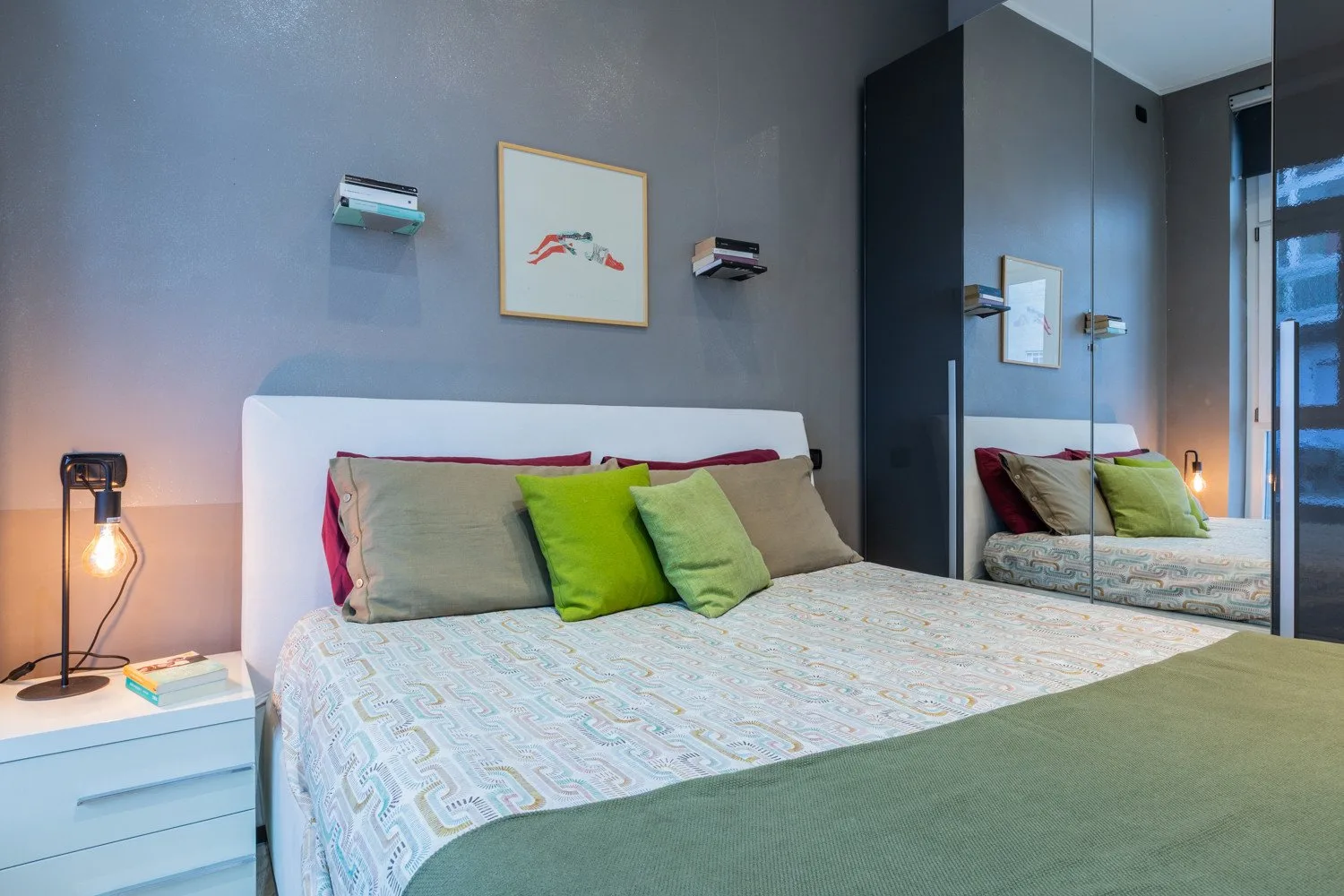 A bedroom with a bed dressed in pale patterned bedding and green cushions.