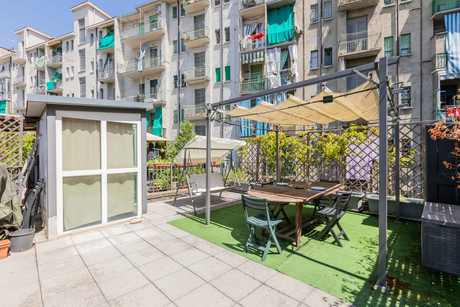 A paved outdoor patio has a shaded seating area beside a small garden.