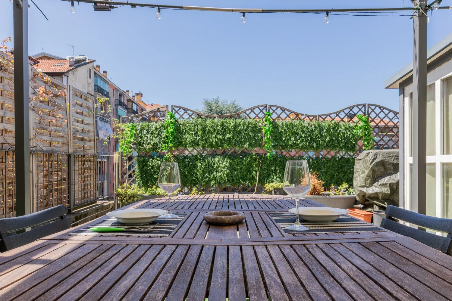 An outdoor dining area with a wooden table beside a leafy trellis.
