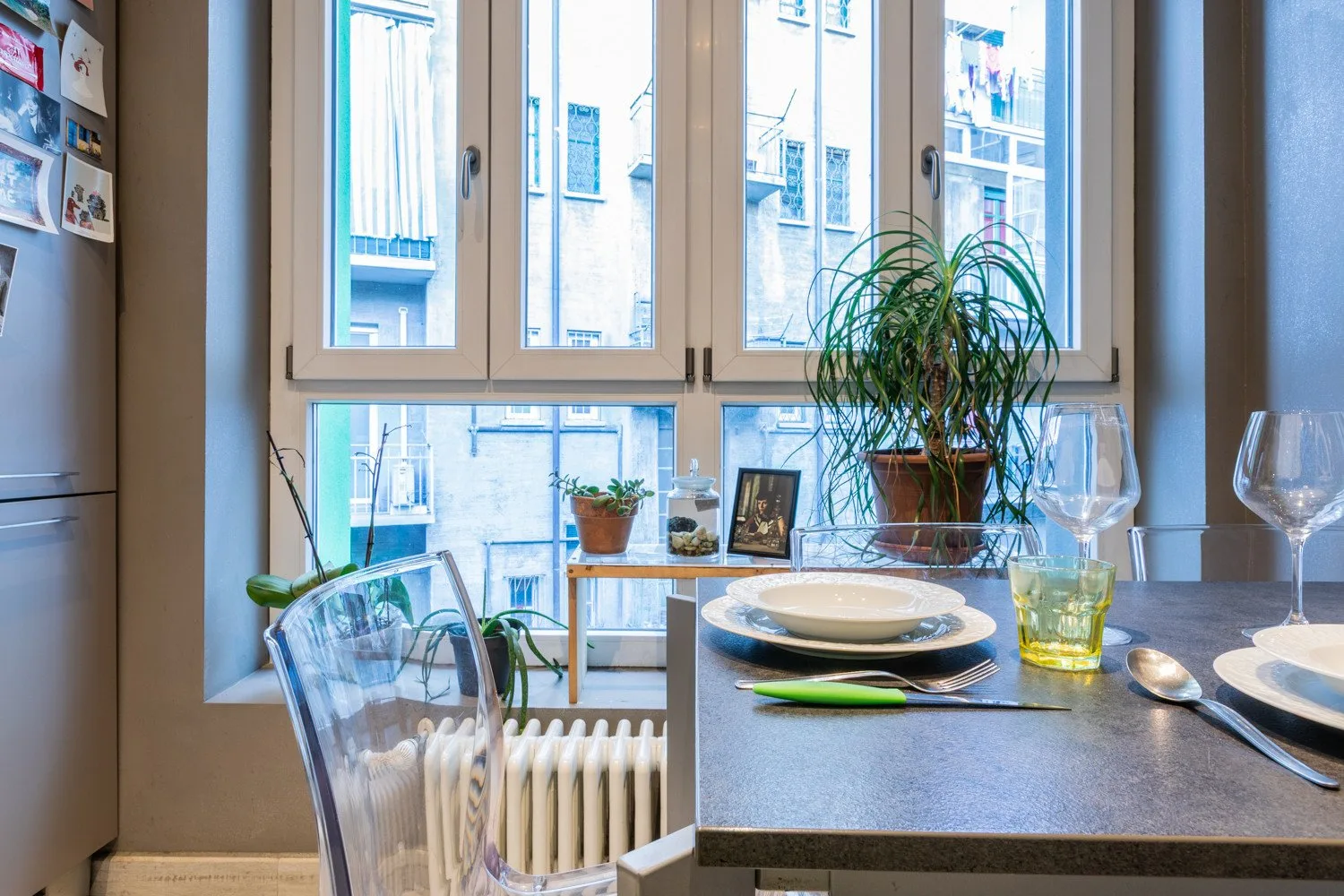 A dining table is set beside large windows, with potted plants on the sill.