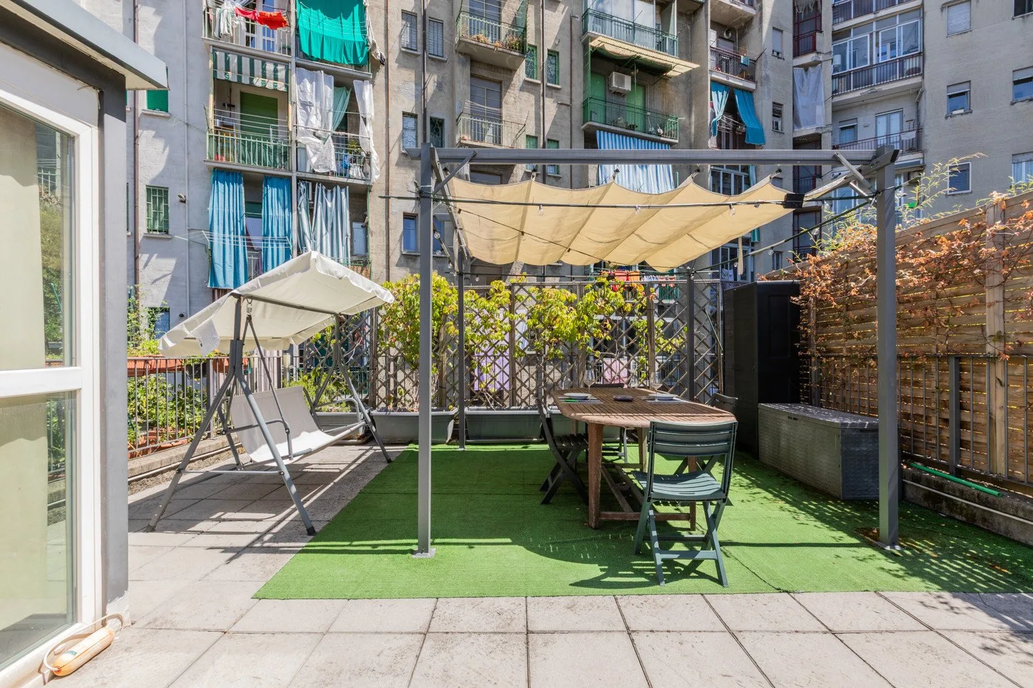 A paved outdoor terrace has a dining table, seating, and a canopy.