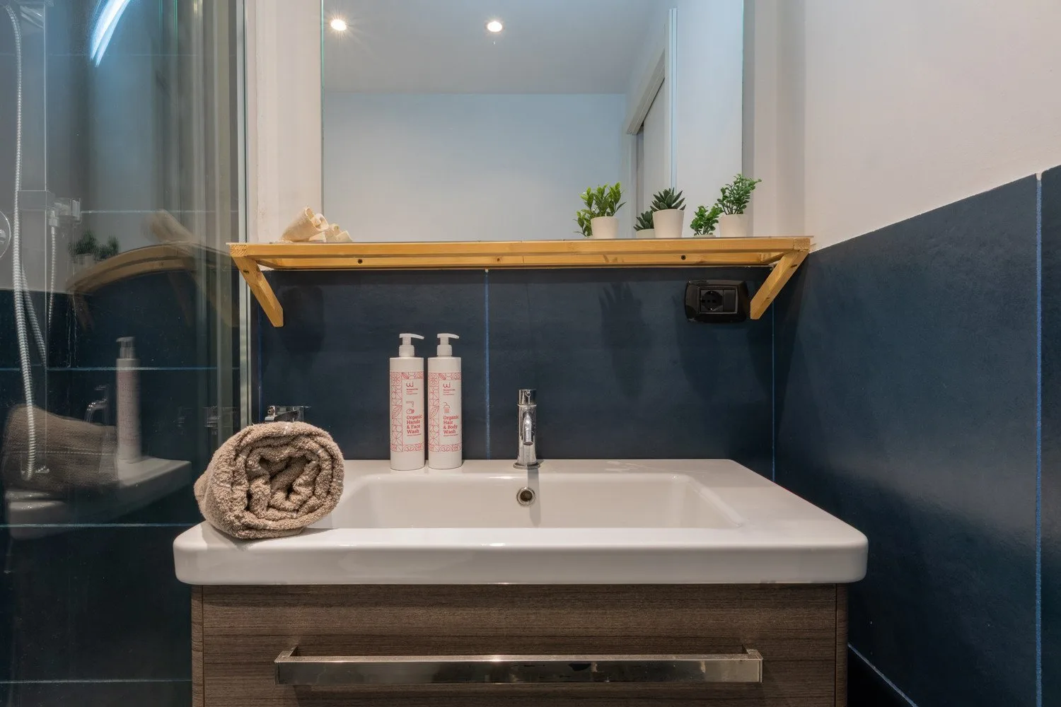 A bathroom vanity with a white sink sits beneath a wooden shelf and mirror.
