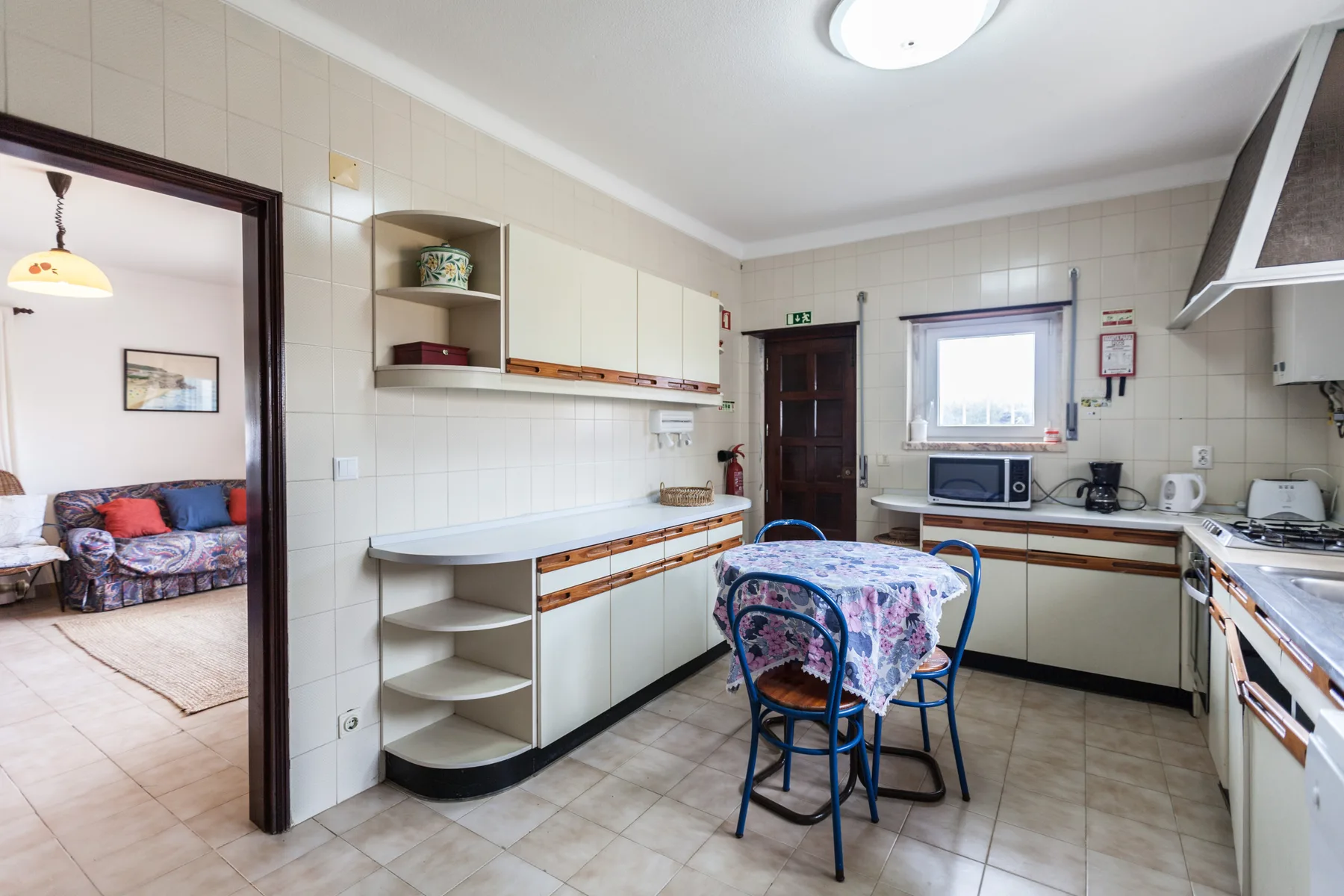 A tiled kitchen with a table and blue chairs, with a sitting area visible through the doorway.
