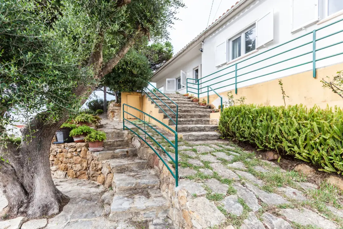 An outdoor stone stairway leads up beside a white building and leafy garden.