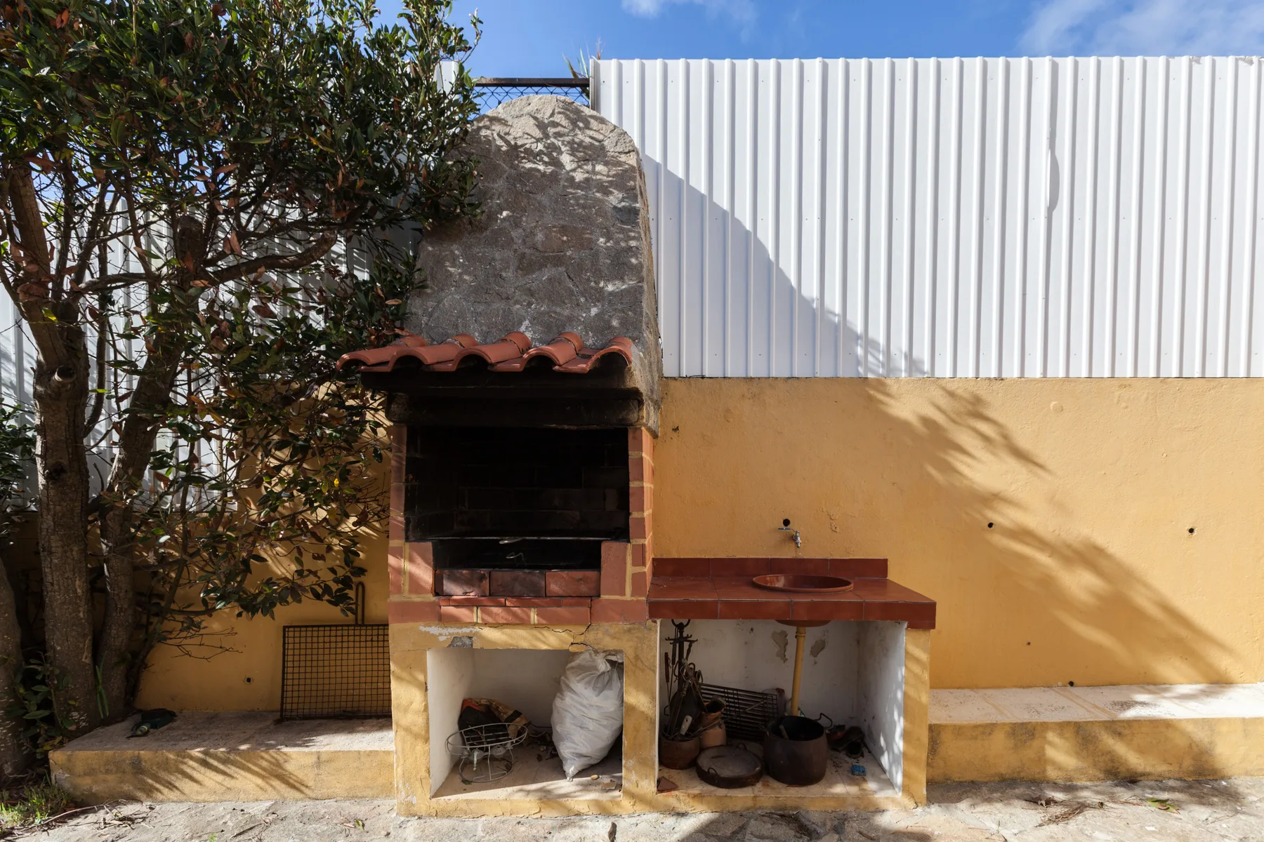 An outdoor brick barbecue stands beside a yellow wall and leafy tree.