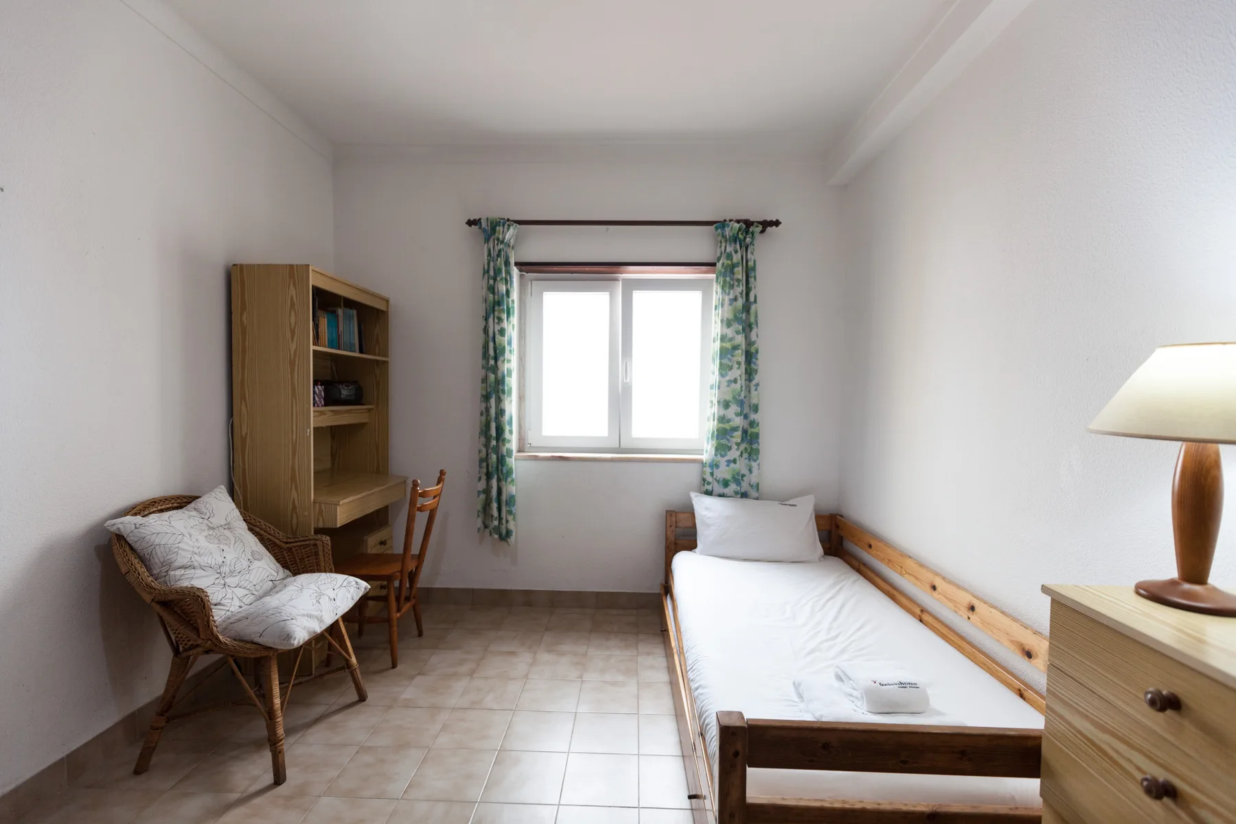 A simple bedroom with a single bed, wooden desk, and window with patterned curtains.