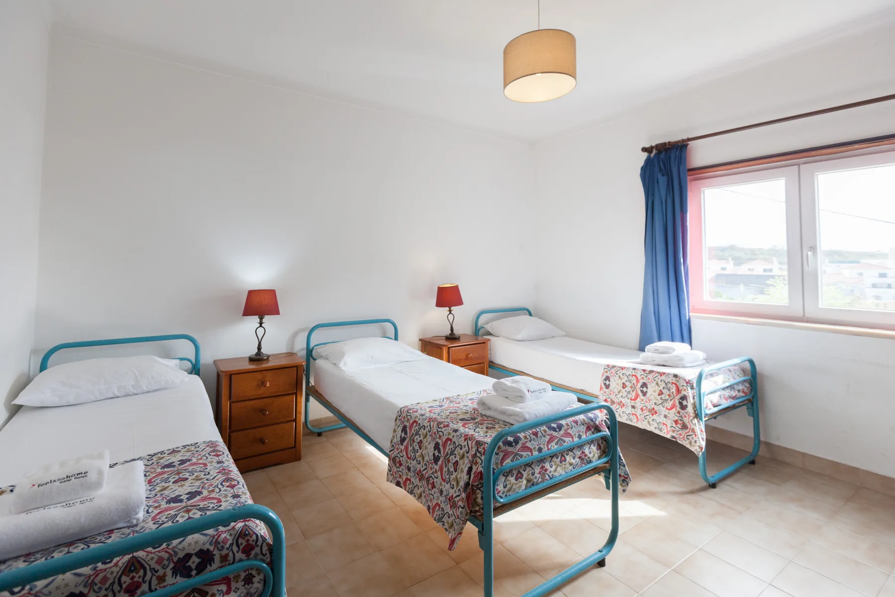 A bright bedroom with three beds and a window framed by blue curtains.
