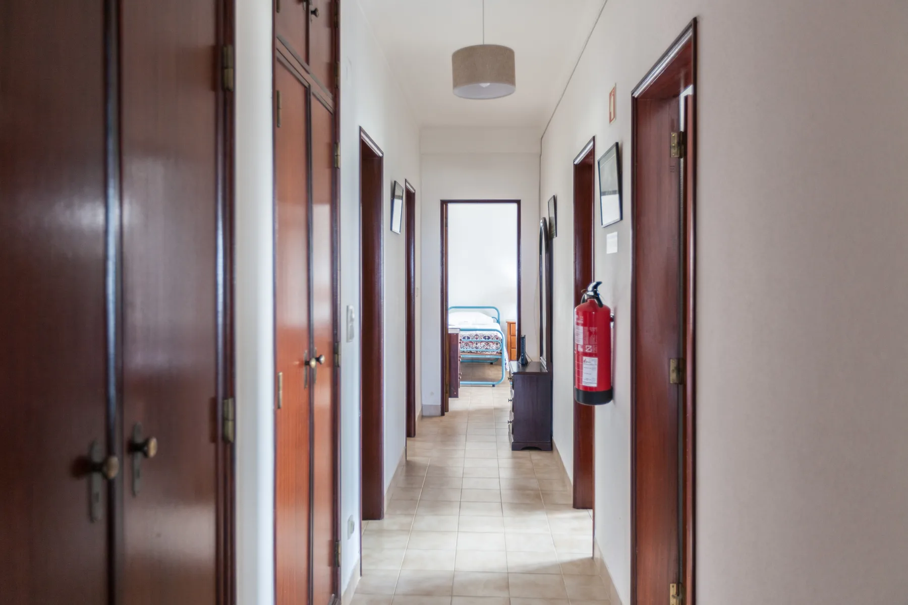 A tiled hallway with several wooden doors leads to a room with a bed.