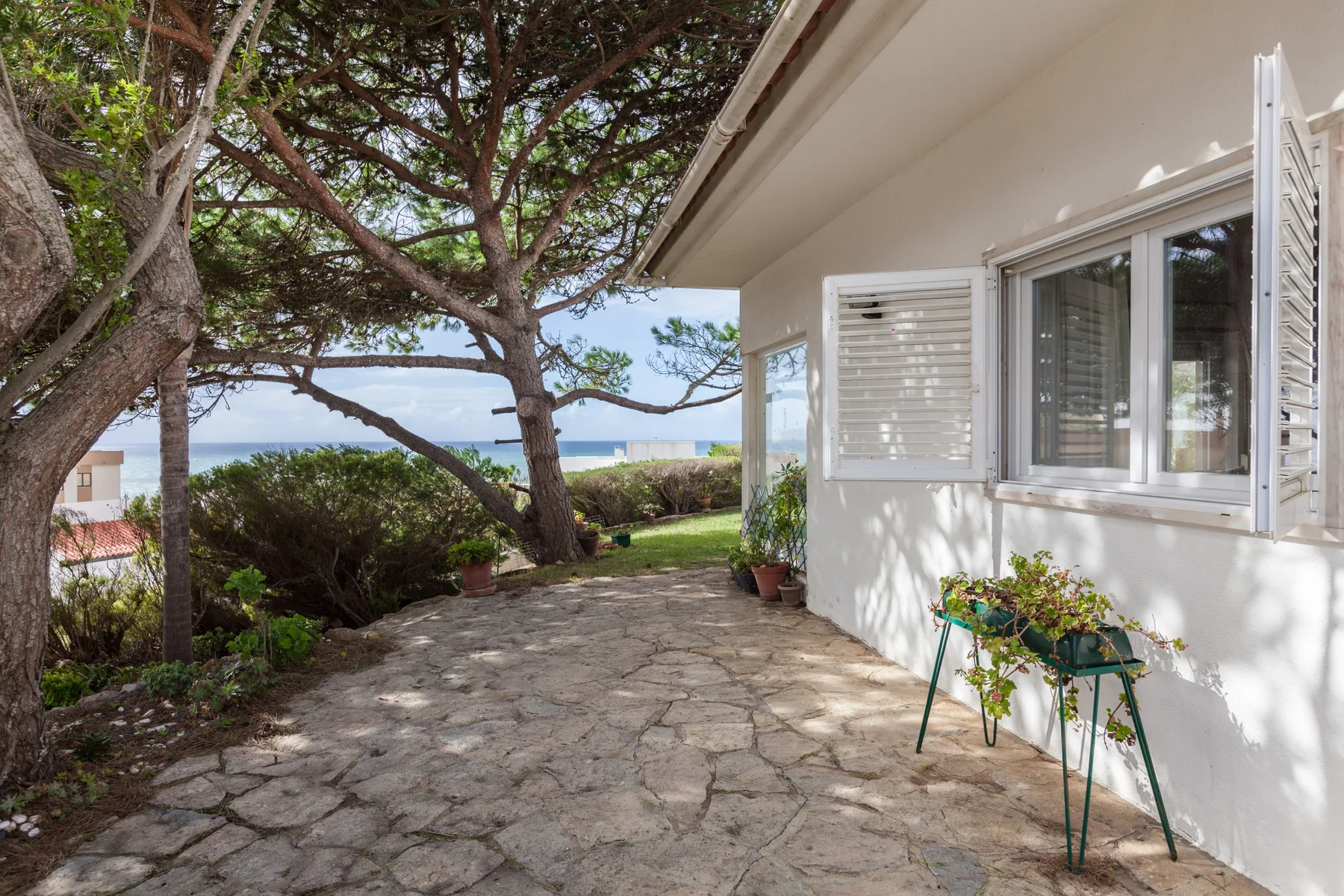A stone-paved patio beside a white house, with trees and the sea beyond.