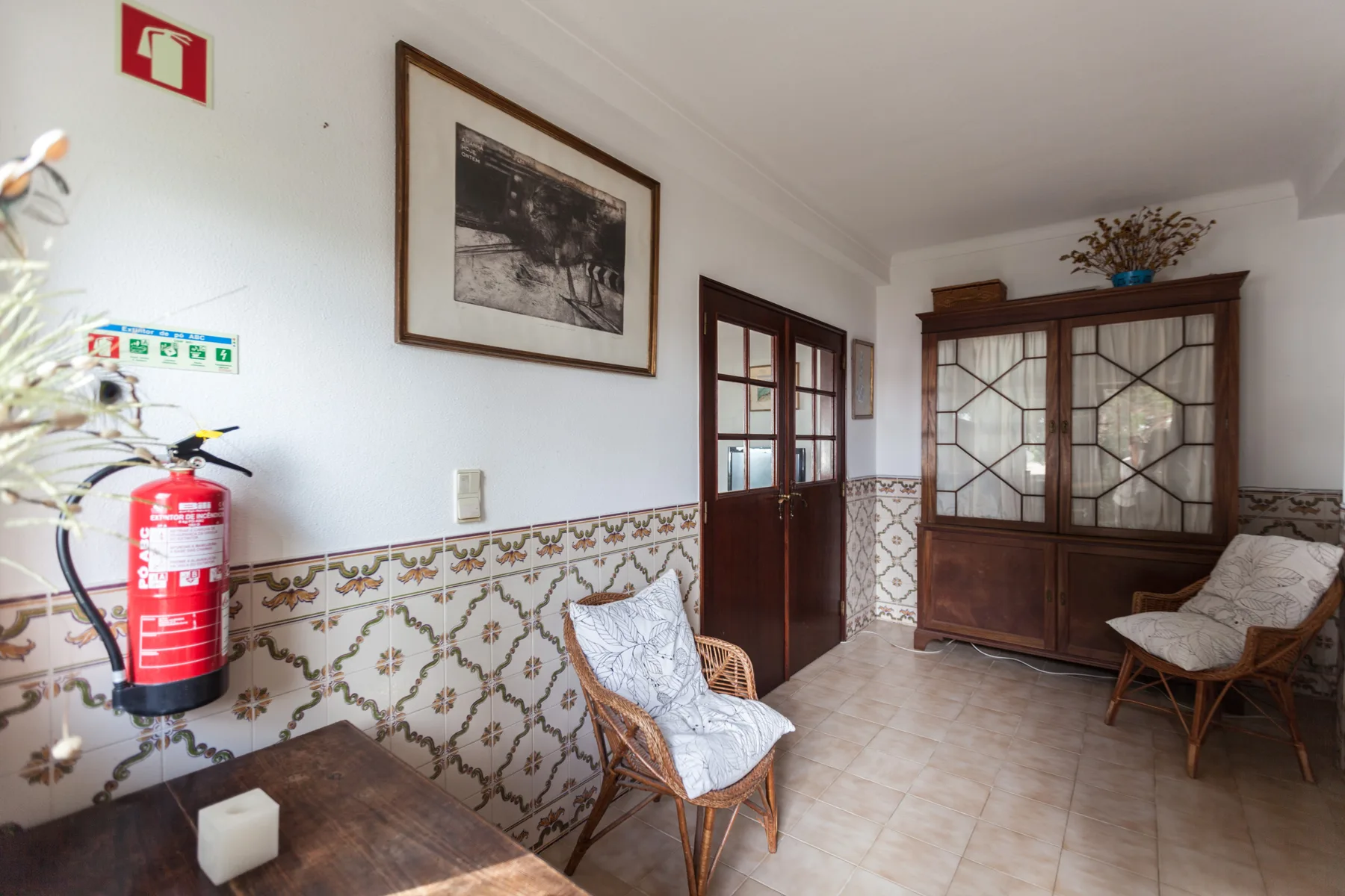 A tiled sitting room with a glass-fronted wooden cabinet and two chairs.